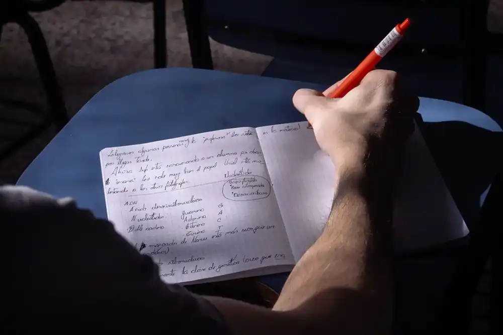Persona escribiendo notas en un cuaderno durante una sesión de estudio, sosteniendo un bolígrafo rojo, en un ambiente de aprendizaje enfocado y silencioso.
