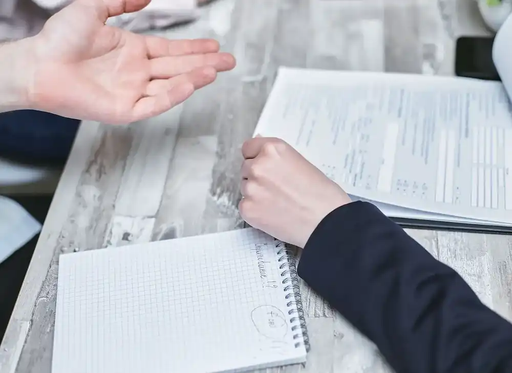Persona explicando un documento a otra durante una conversación de trabajo, con cuaderno abierto sobre la mesa y laptop al fondo, representando planificación y formación profesional.