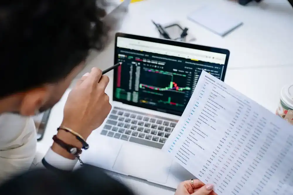 Persona revisando gráficos de mercado y métricas en una laptop mientras analiza un reporte financiero en un entorno de oficina.