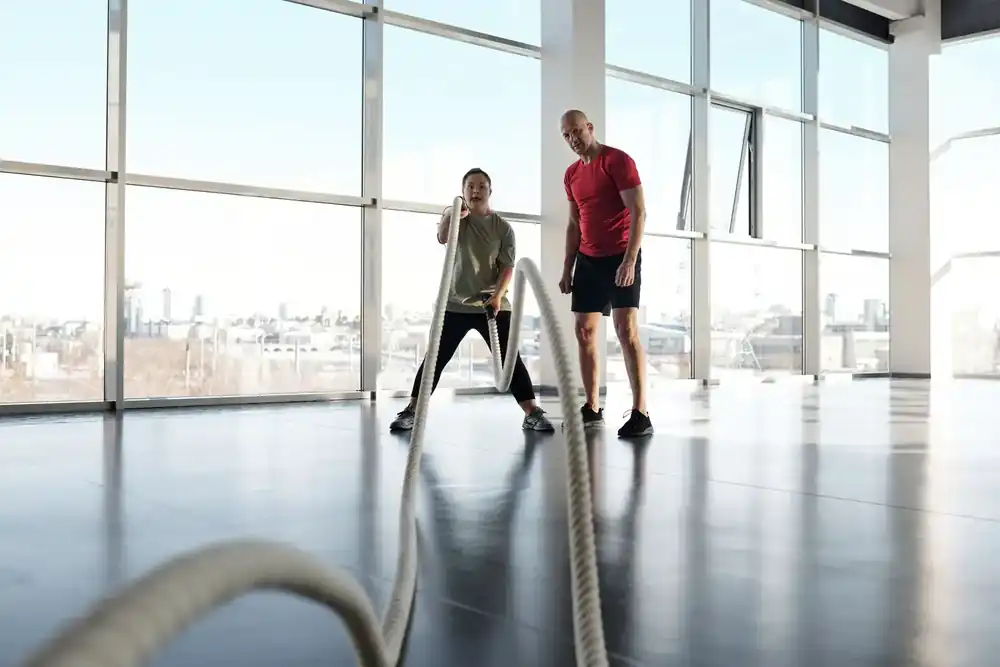 Entrenador acompaña a una socia durante un ejercicio con cuerdas en un gimnasio amplio y luminoso.